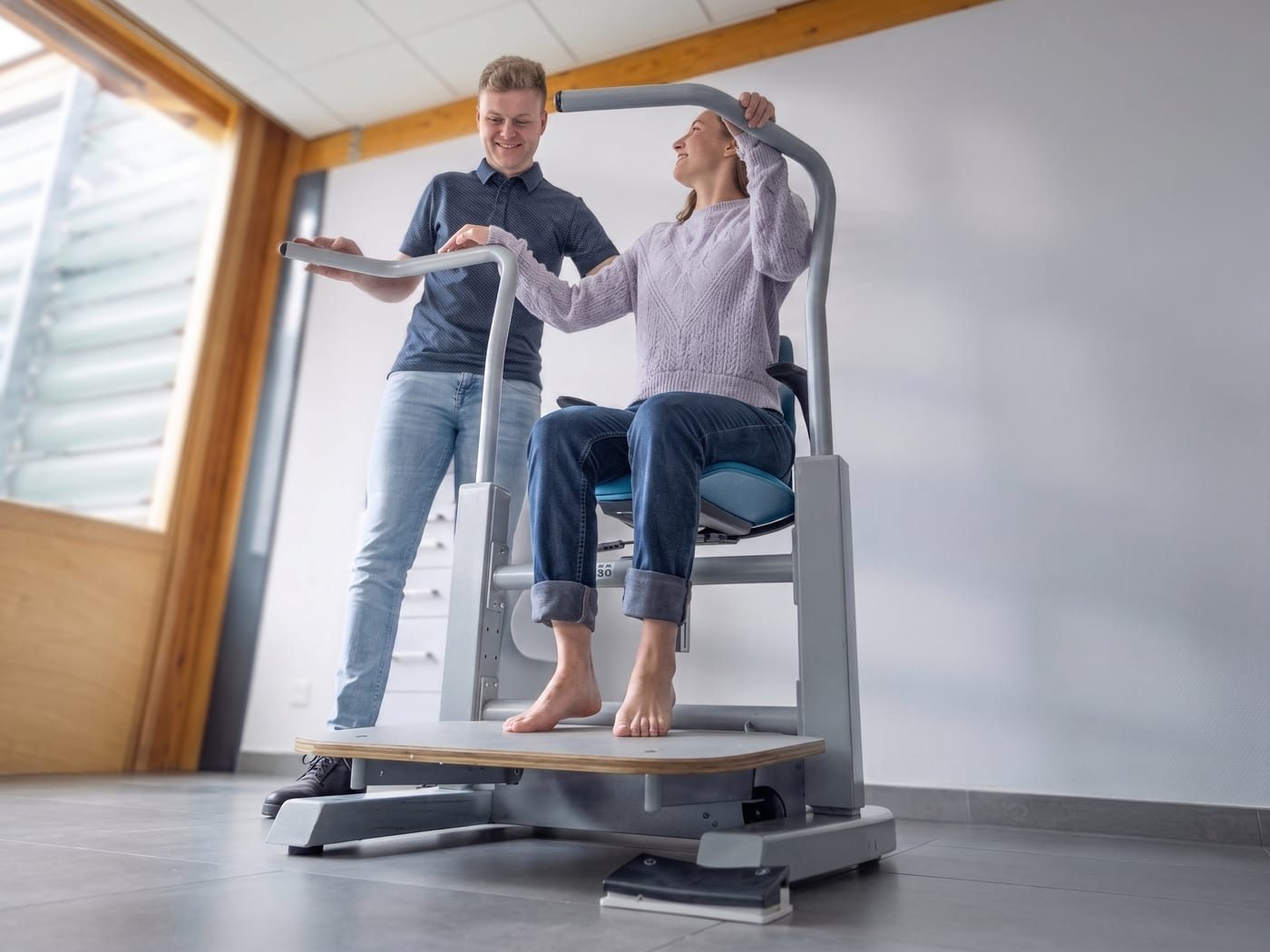Eine Klientin sitzt auf dem angehobenen Orthostand-Sitz und hält sich an der Stehstütze fest, während die Fachkraft aufrecht neben ihr arbeitet