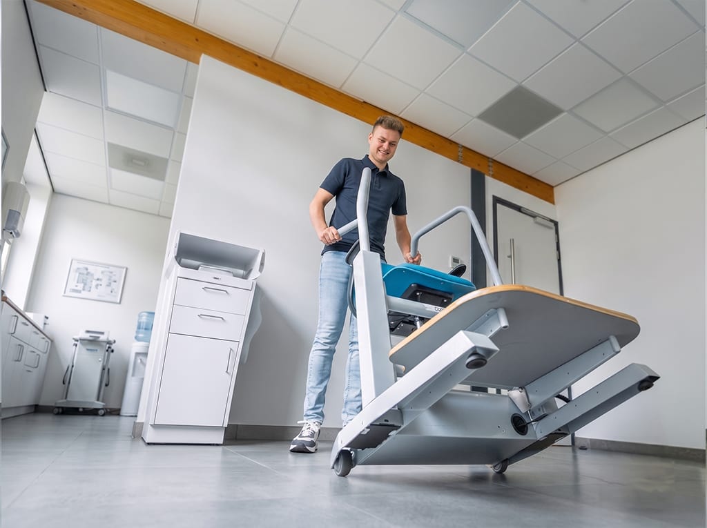 Eine Fachkraft rollt den Orthostand M30 auf seinen Rollen durch einen Behandlungsraum, die Plattform in die Transportstellung gekippt