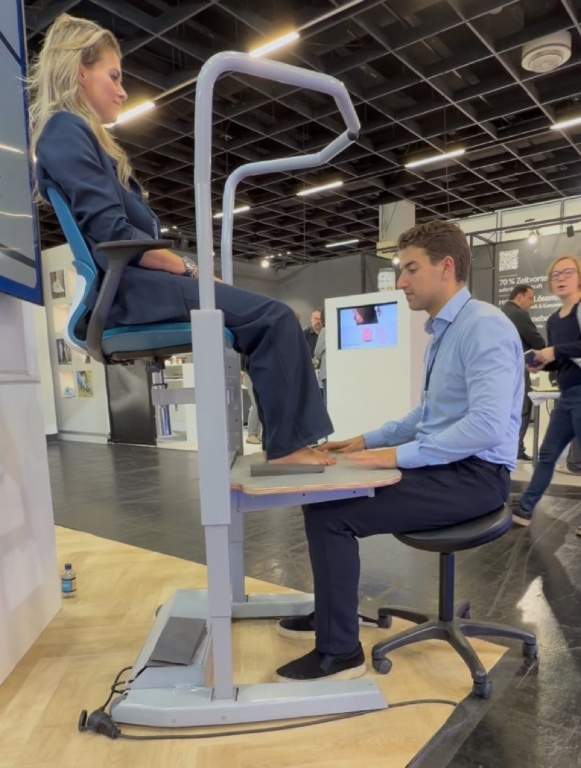 A specialist sitting upright on a stool, treating a client whose foot is raised to working height on the Orthostand, assessed under load